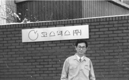 Black-and-white photo of Chairman Kyung-Soo Lee in uniform under COSMAX sign on brick wall
