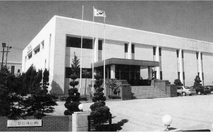 Black-and-white exterior of COSMAX Hwaseong Plant 2 with Korean flag on left roof and COSMAX sign on brick wall
