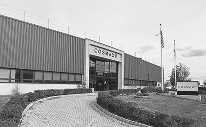 Black-and-white COSMAX USA factory with US and Korean flags, white sign with COSMAX logo