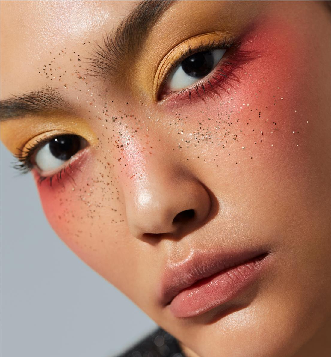 Model tilting head about 15 degrees left with yellow-toned makeup above eyes, pink tone below, and glitter accents
