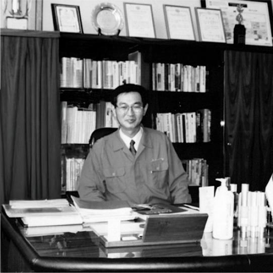 Cosmax Group Chairman Lee Kyungsoo in office uniform looking at camera, bookshelf with books in background, desk with documents and cosmetics
